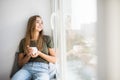 Beautiful young woman drinking coffee and looking through window while sitting at windowsill at home Royalty Free Stock Photo