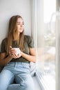 Beautiful young woman drinking coffee and looking through window while sitting at windowsill at home Royalty Free Stock Photo