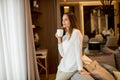 Beautiful young woman drinking coffee and looking through window while standing in the apartment Royalty Free Stock Photo