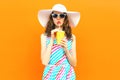 Beautiful young woman drinking juice in summer straw hat, colorful striped dress on orange wall