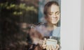 Beautiful young woman drinking morning tea or coffee while sitting by the window in her country house, in the glass Royalty Free Stock Photo