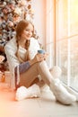 Beautiful young woman drinking tea at the Christmas tree. Beautiful girl celebrates Christmas with cup of cocoa.