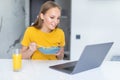 Beautiful young woman eat breakfast leaning on the kitchen table with laptop Royalty Free Stock Photo