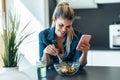 Beautiful young woman eating a bowl of salad while using her mobile phone in the kitchen at home Royalty Free Stock Photo