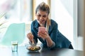 Beautiful young woman eating a bowl of salad while using her mobile phone in the kitchen at home Royalty Free Stock Photo