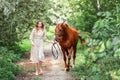 Beautiful young woman in elegant lifestyle dress walking together with her friend red horse in green forest