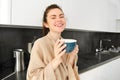 Beautiful young woman enjoys her morning mug of coffee, drinking tea from cup in the kitchen, wearing bathrobe and Royalty Free Stock Photo