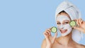 Beautiful young woman with facial mask on her face holding slices of cucumber. Skin care and treatment, spa, natural beauty and