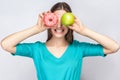 Beautiful young woman with freckles in green dress, holding before her eyes green apple and pink donut and shows her teeth.