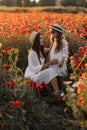 Beautiful young woman with girl in field with poppies, mother and daughter in white dresses and straw hats in evening at sunset,