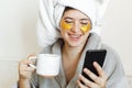 Beautiful young woman with golden eye patches in bathrobe holding cup of coffee, lying in bed and looking at phone screen. Royalty Free Stock Photo