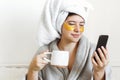 Beautiful young woman with golden eye patches in bathrobe holding cup of coffee, lying in bed and looking at phone screen. Royalty Free Stock Photo