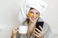 Beautiful young woman with golden eye patches in bathrobe holding cup of coffee, lying in bed and looking at phone screen. Royalty Free Stock Photo