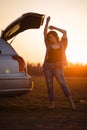 Beautiful young woman happy and dancing in a car`s trunk during a road trip in Europe in the last minutes of Golden Hour Royalty Free Stock Photo
