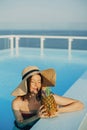 Beautiful young woman in hat drinking delicious cocktail from pineapple and relaxing in pool, summer vacation. Girl enjoying warm Royalty Free Stock Photo