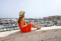 Beautiful young woman with hat sitting on wall looking the port of Bisceglie, Apulia, Italy