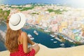 Beautiful young woman with hat sitting on wall looking at stunning panoramic view of Procida Island, Naples, Italy