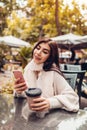 Beautiful young woman having coffee in outdoor cafe while using smartphone. Portrait of stylish girl typing message Royalty Free Stock Photo