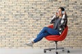 Beautiful young woman with headphones listening to music while sitting on armchair near brick wall Royalty Free Stock Photo