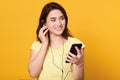 Beautiful young woman in headphones listening to music via her modern mobile phone, smiling lady standing over yellow background Royalty Free Stock Photo