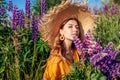 Beautiful young woman holding bouquet of purple lupin flowers walking in summer meadow. Stylish girl wearing straw hat Royalty Free Stock Photo