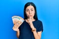 Beautiful young woman holding bunch of 50 euro banknotes asking to be quiet with finger on lips