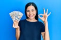 Beautiful young woman holding dollars smiling positive doing ok sign with hand and fingers Royalty Free Stock Photo