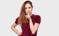 Beautiful young woman holding her index finger to her lips to keep silence, on white wall. Pretty female wears red t-shirt, Royalty Free Stock Photo