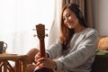 A beautiful young woman holding and looking at an ukulele while enjoy listening to music with headphone at home