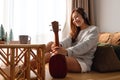 A beautiful young woman holding and looking at an ukulele while enjoy listening to music with headphone at home