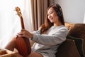 A beautiful young woman holding and looking at an ukulele while enjoy listening to music with headphone at home