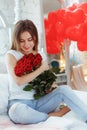 Beautiful young woman at home. Attractive girl is sitting on bed with air baloons in shape of heart and red roses. Celebrating Royalty Free Stock Photo