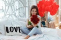 Beautiful young woman at home. Attractive girl is sitting on bed with gift box, air baloons in shape of heart and red roses. Royalty Free Stock Photo