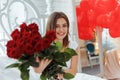 Beautiful young woman at home. Attractive girl is sitting on bed with gift box, air in shape of heart and red roses. Celebrating Royalty Free Stock Photo