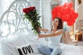 Beautiful young woman at home. Attractive girl sitting on bed with red roses and gift box, heart shaped balloons. Valentine`s Day Royalty Free Stock Photo