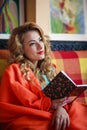 Beautiful young woman at home on sofa wrapped in a warm blanket in the room reading a book dreamily Royalty Free Stock Photo