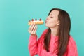 Beautiful young woman in knitted pink sweater with closed eyes holding in hand eating blowing sending air kiss to eclair