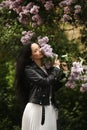 Beautiful young woman in leather jacket enjoying the smell of the bloomy lilac tree in springtime