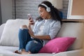 Beautiful young woman listening to music with headphones while using her smartphone sitting on sofa at home Royalty Free Stock Photo