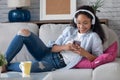 Beautiful young woman listening to music with headphones while using her smartphone sitting on sofa at home Royalty Free Stock Photo