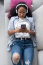 Beautiful young woman listening to music with headphones while using her smartphone sitting on sofa at home Royalty Free Stock Photo