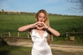 beautiful young woman with long blonde hair in white dress smiling in love making the shape of a heart symbol with her hands. Royalty Free Stock Photo