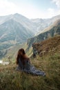 Beautiful young woman in a long dress is sitting on a cliff in the background of mountains