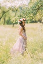 Beautiful young woman in long lilac dress with wreath on head standing back outdoors