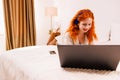 Beautiful young woman with long red hair and white shirt lying on her stomach in bed enjoying watching something on her Royalty Free Stock Photo