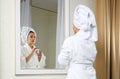 Beautiful young woman look in mirror applying cream, pretty lady wrap towel on head put facial moisturizer lifting Royalty Free Stock Photo