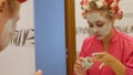 Beautiful young woman looks in the mirror and applies a cosmetic face mask. A pretty blonde in curlers on her head is Royalty Free Stock Photo