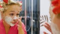 Beautiful young woman looks in the mirror and applies a cosmetic face mask. A pretty blonde in curlers on her head is Royalty Free Stock Photo