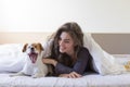 Beautiful young woman lying on bed with her cute small dog besides and yawning. Home, indoors and lifestyle Royalty Free Stock Photo