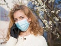 Beautiful young woman in a medical mask on the background of a blossoming spring tree speaks by phone